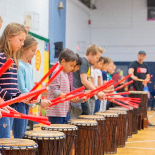 Children playing drums in a line with red drumsticks in an indoor setting